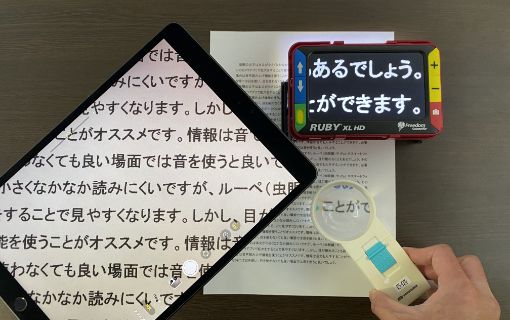 A４サイズの紙にルーペやタブレット端末、拡大読書器を置いて文字を大きく表示している写真